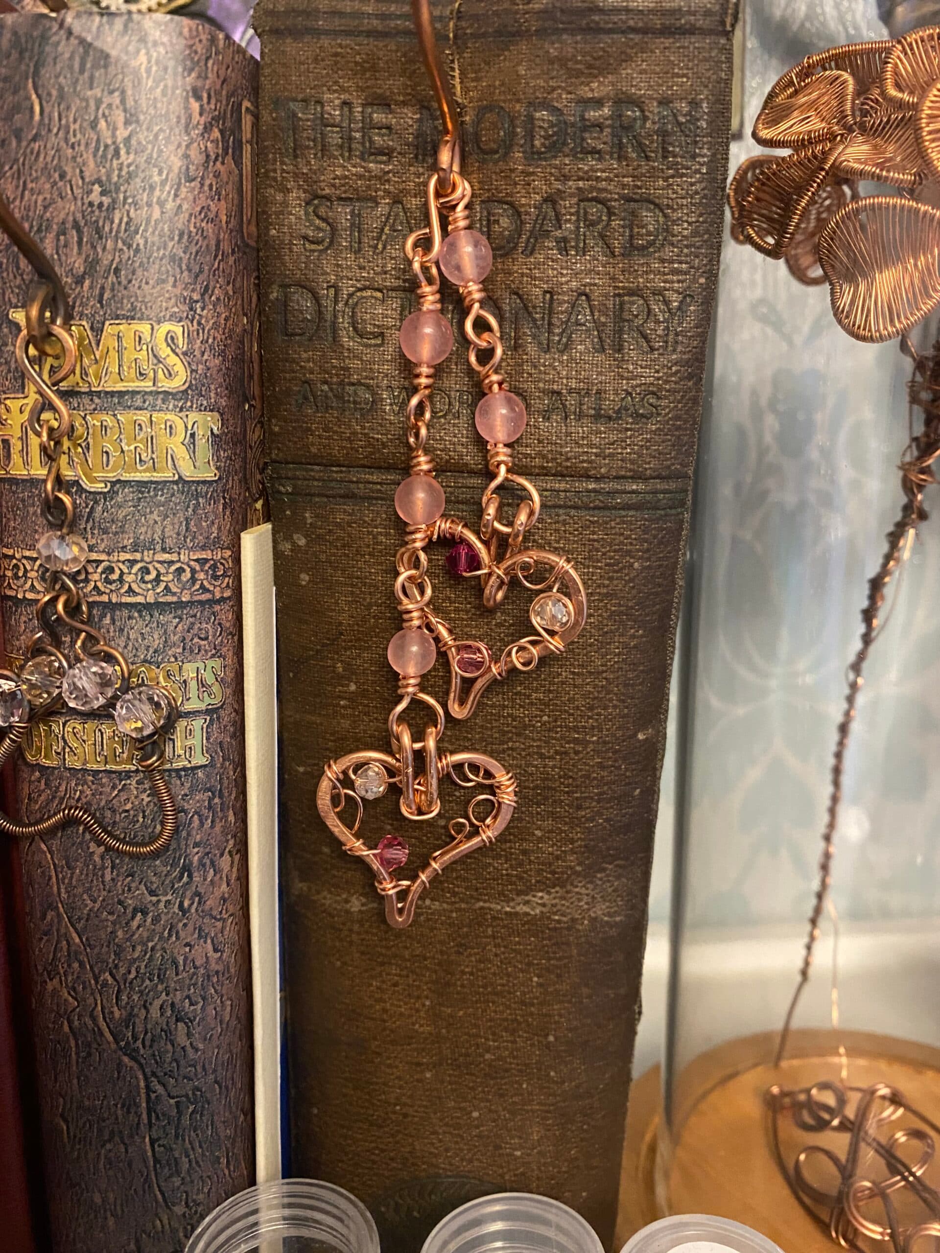 Handmade copper bookmark with two hearts and pink beads hanging on shepherd’s crook chain over hardback book spine