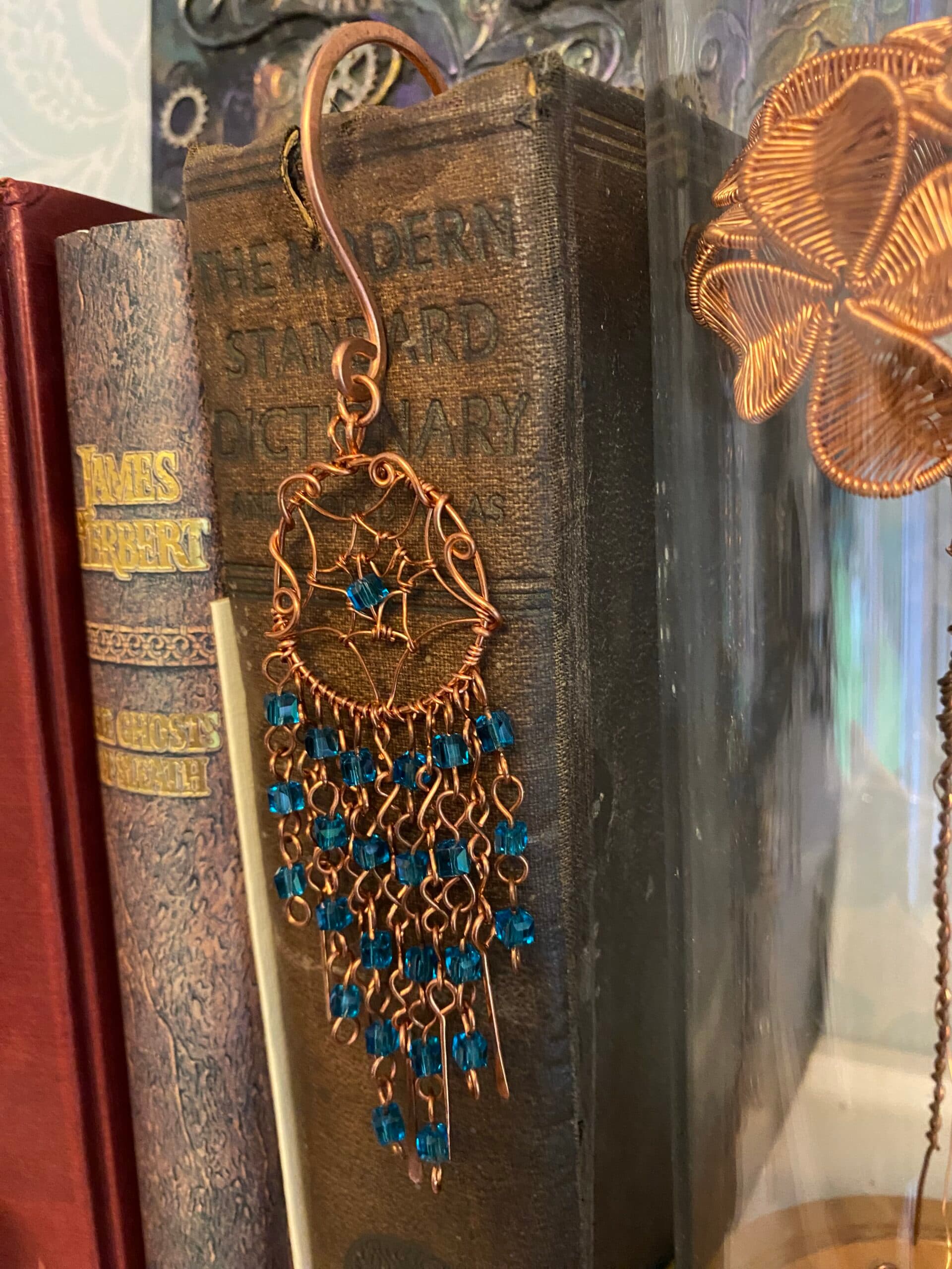 Handmade copper dreamcatcher bookmark with light blue beads, beaded V-shaped tassels, and hammered copper paddles hanging on hardback book spine