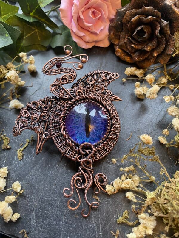Handmade copper dragon eye brooch with purple and blue hand-painted eye, ornate wire woven wings and flowing copper scrolls.