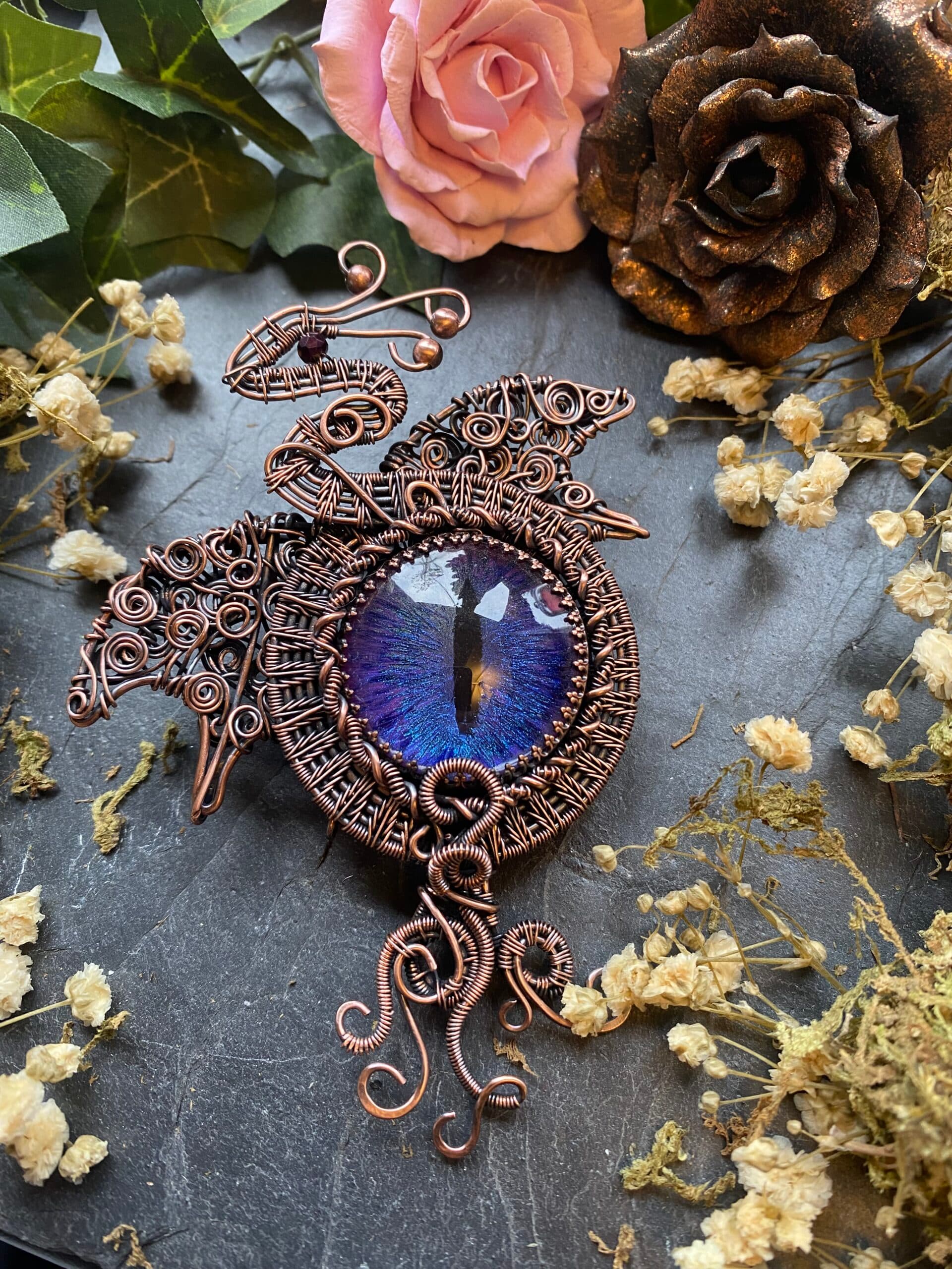 Handmade copper dragon eye brooch with purple and blue hand-painted eye, ornate wire woven wings and flowing copper scrolls.