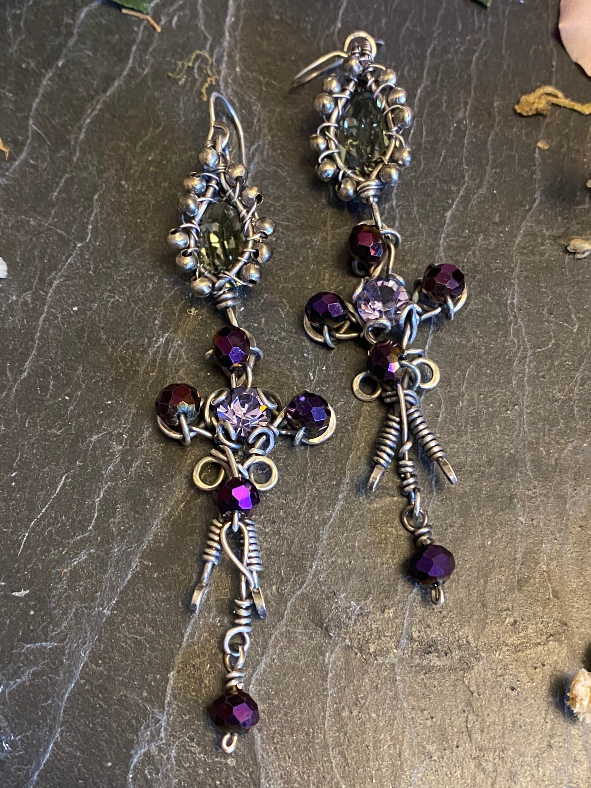 Pair of handmade gothic earrings featuring antiqued silver filled wire, purple Swarovski crystals and long decorative wire-wrapped drops on dark slate.