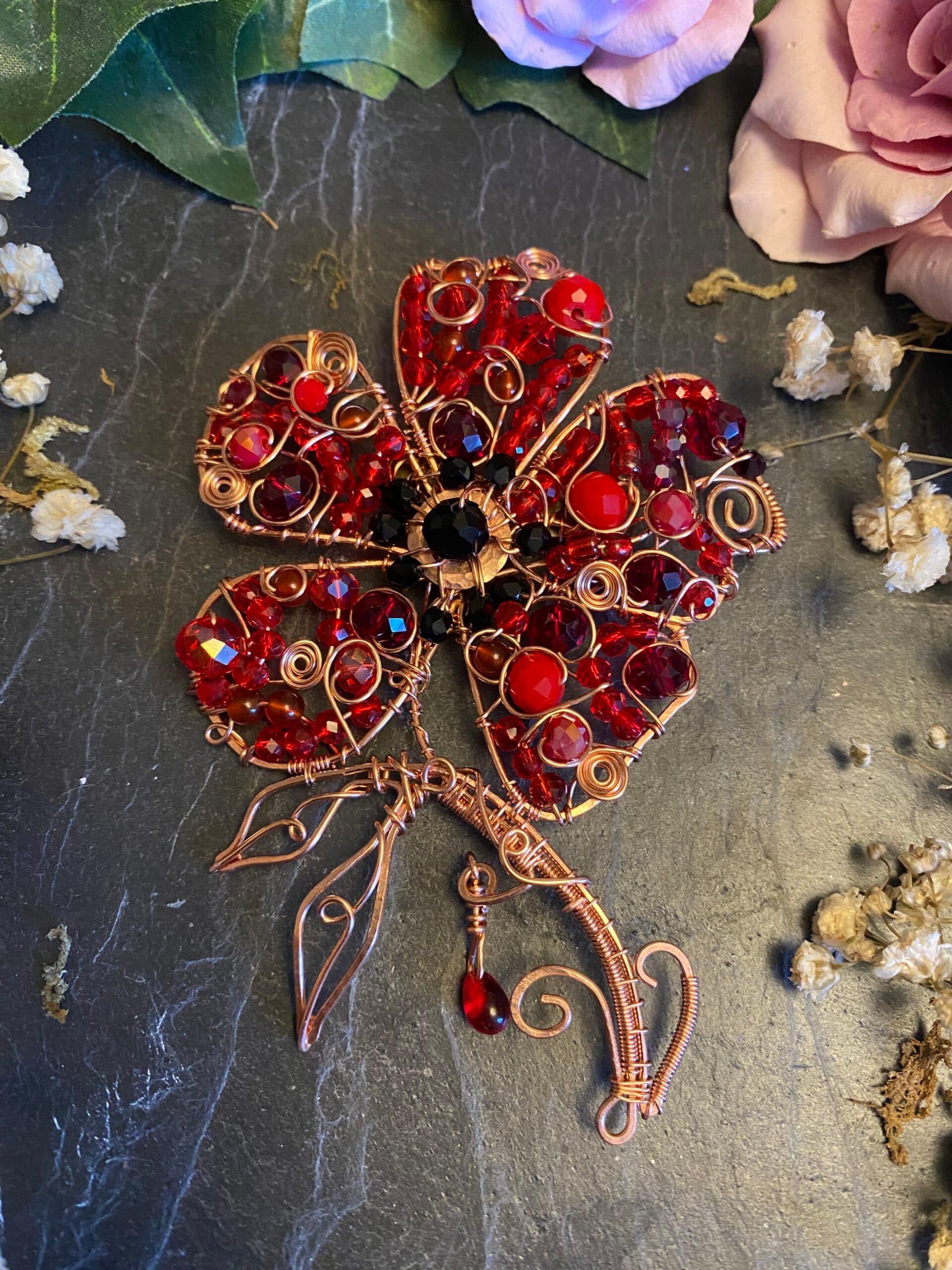 Handmade copper wire Remembrance poppy brooch with five red beaded petals, black centre, copper leaves and a symbolic dangling red bead, photographed on dark slate.