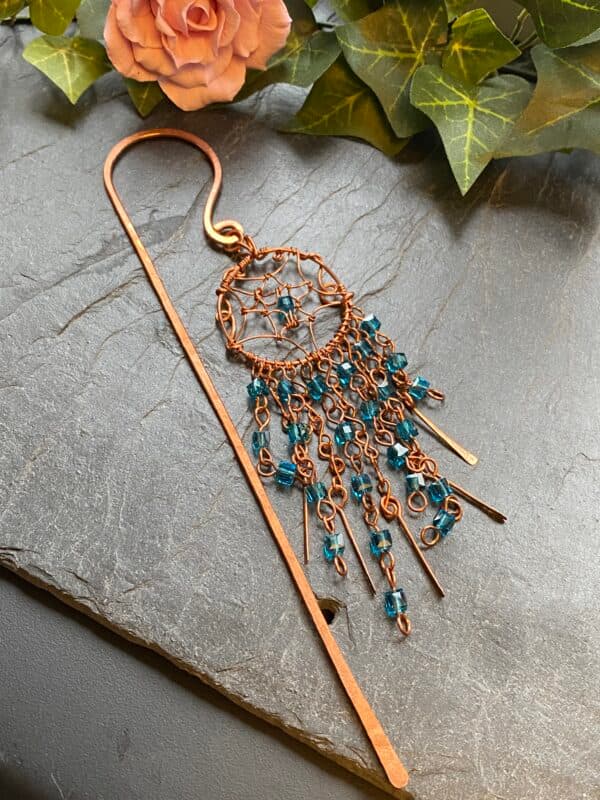 Handmade copper dreamcatcher bookmark with light blue beads, beaded V-shaped tassels, and hammered copper paddles hanging on hardback book spine