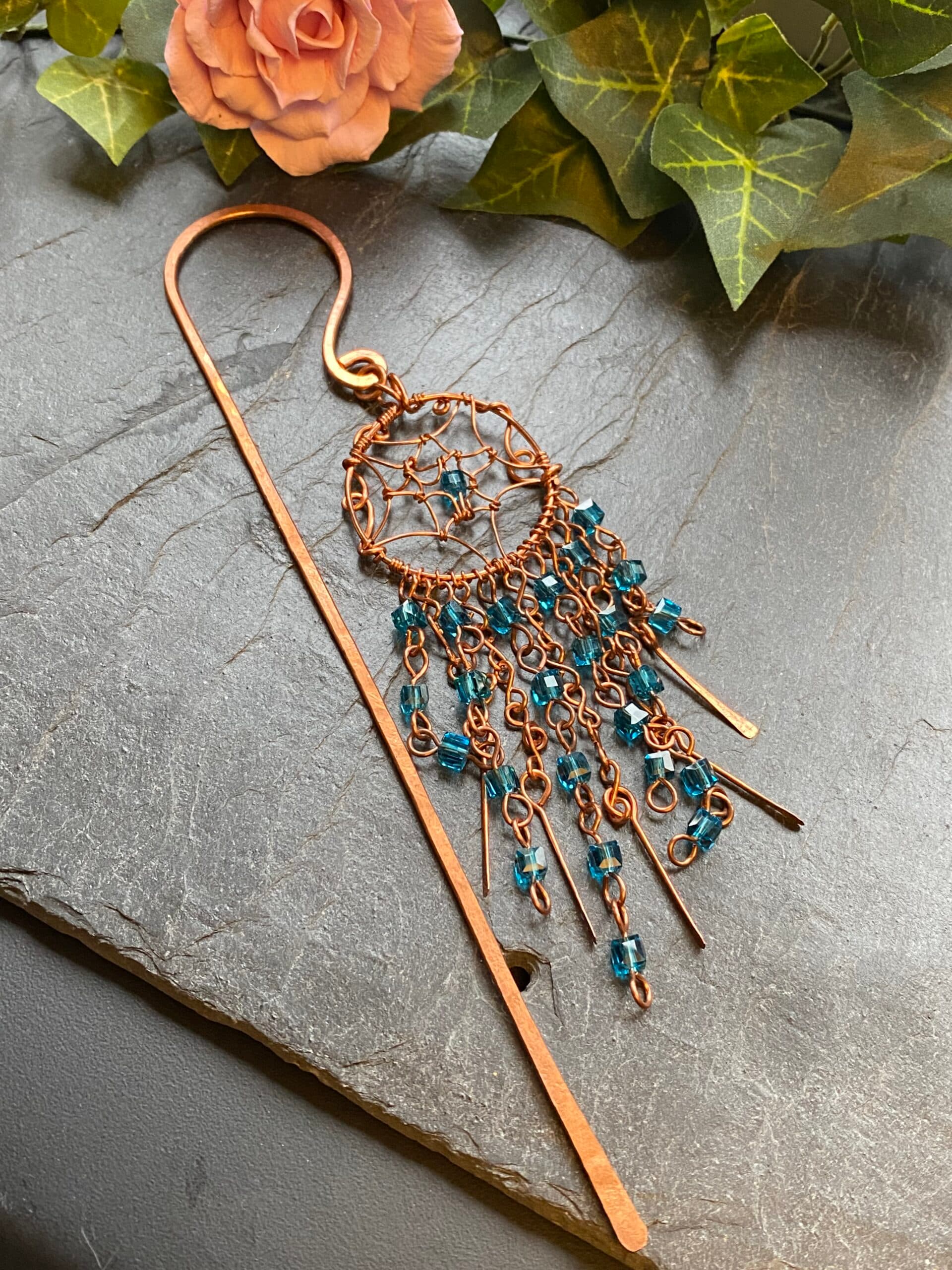 Handmade copper dreamcatcher bookmark with light blue beads, beaded V-shaped tassels, and hammered copper paddles hanging on hardback book spine