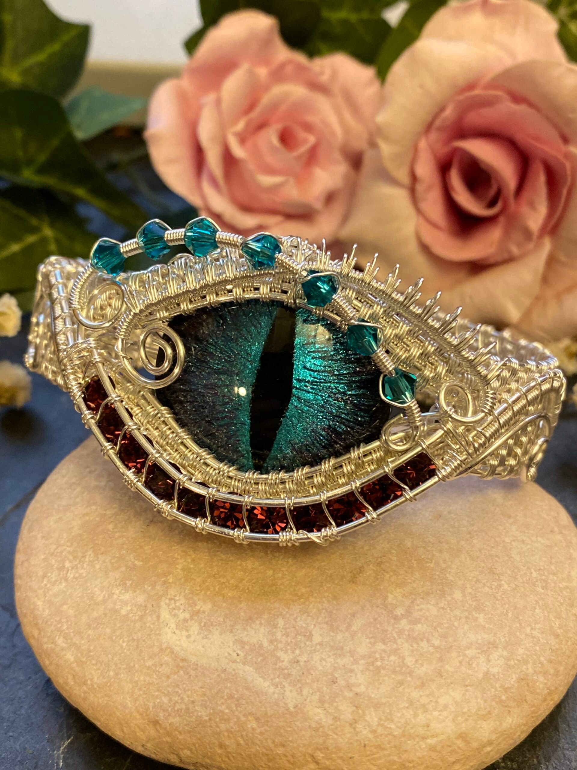 Handmade silver filled dragon eye cuff bracelet displayed on a stone, with teal Swarovski bicone beads and purple crystals framing the eye.