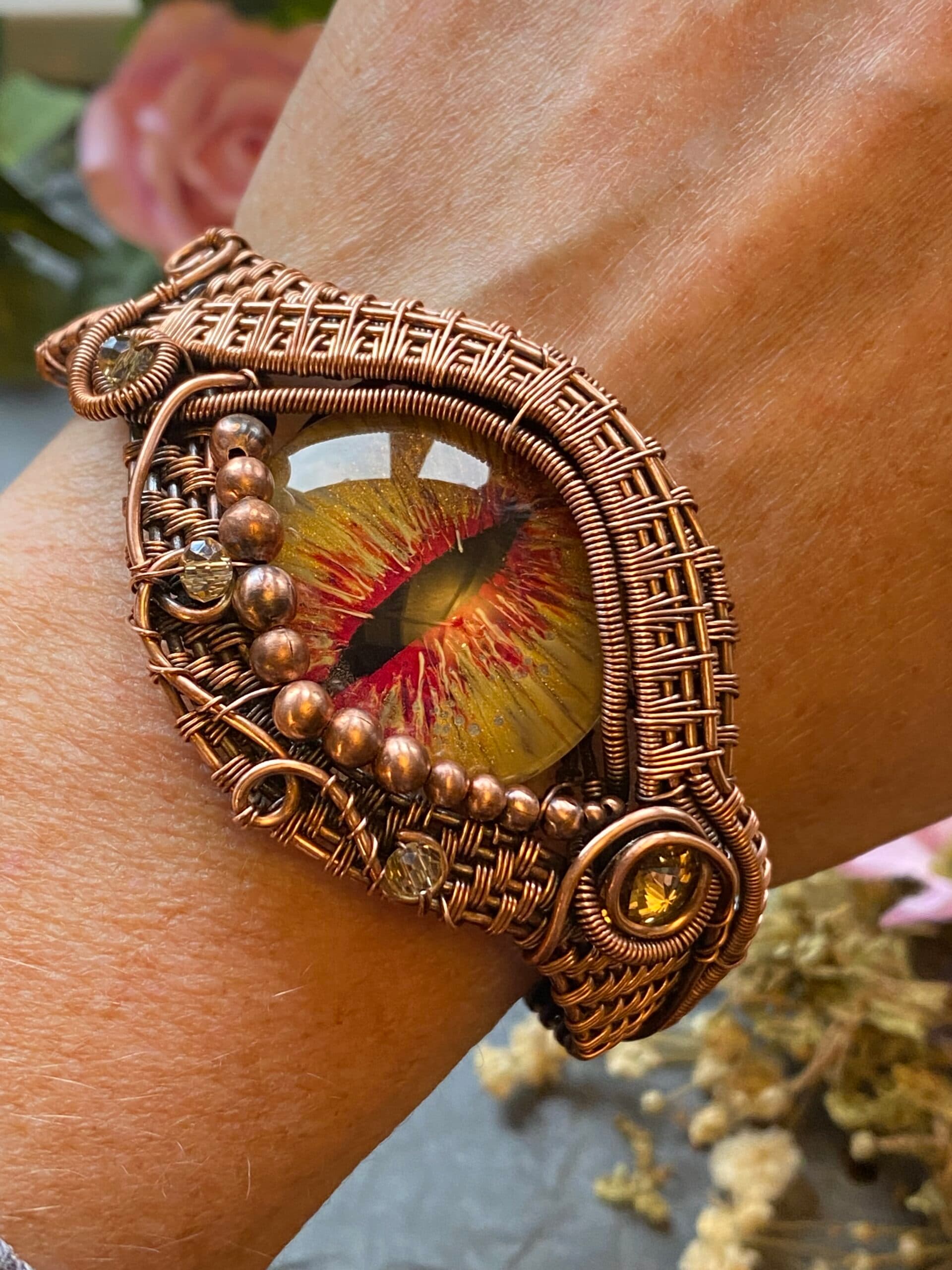 Yellow and red hand-painted dragon eye cuff bracelet worn on a wrist, showing detailed copper wire weaving and decorative beads